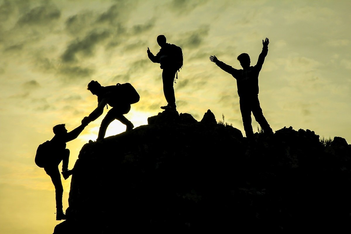 Silhouettes of four hikers on a mountain at sunset, with one person helping another climb, balancing teamwork, support, and leadership.